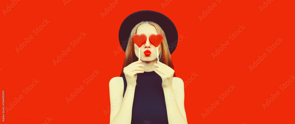 Portrait of beautiful young woman with lollipop blowing her lips with lipstick sending sweet air kiss wearing red heart shaped sunglasses, black round hat on background Portrait of beautiful young woman with lollipop blowing her lips with lipstick sending sweet air kiss wearing red heart shaped sunglasses, black round hat on background