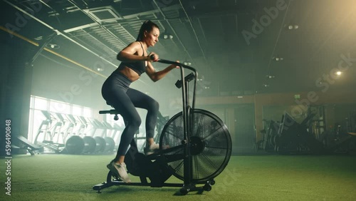 Young well-built cute woman in process of training on air bike in fully equipped gym. Woman with pony tail, sweaty body wears sports top and leggings. High quality 4k footage