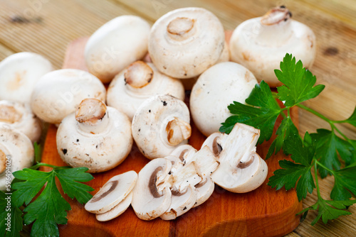 Raw white champignons mushrooms on wooden surface with greens in kitchen