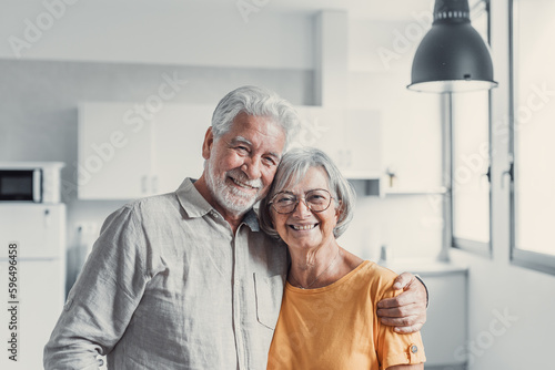 Headshot portrait of smiling elderly 60s husband and wife sit relax on couch hugging cuddling, happy mature old couple rest on sofa in living room embrace look at camera show love and care.