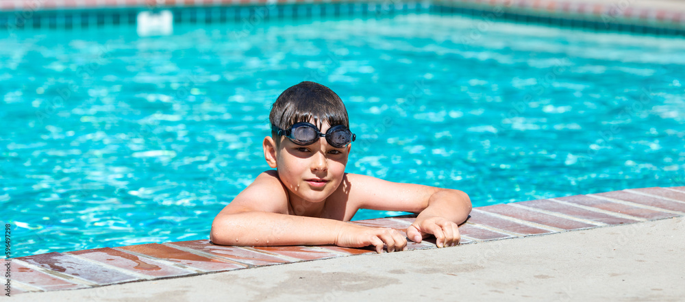 Oudoor summer activity. Concept of fun, health and vacation. A happy boy eight years old in swimming goggles is holding onto the side of the pool on a hot summer day.