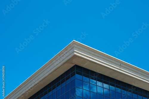 The corner of a futuristic commercial business building. The window panes are blue and reflect the clouds and sky. The eave of the building is cream colored concrete with blue sky in the background