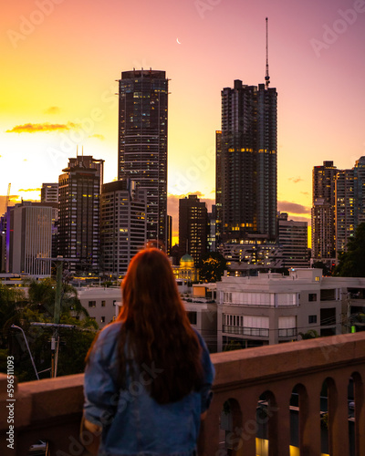 beautiful woman in a hat watching the sunset over brisbane city from kangaroo point, city reach boardwalk with amazing view of large skyscrapers by brisbane river, australia, queensland