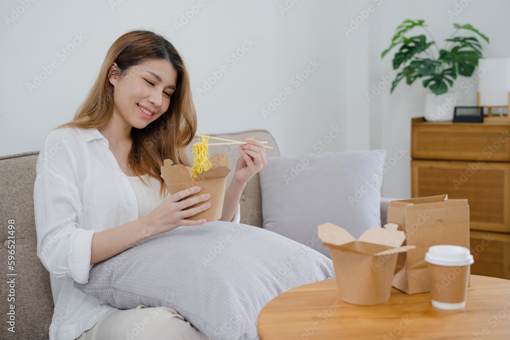 Food delivery. Happy asian woman eating noodles with chopsticks unpacking takeaway food at home.