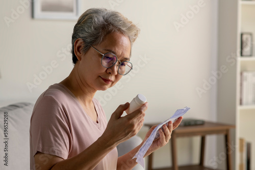 Asian senior woman sitting on sofa in her living room reading the information sheet of her prescribed medicine