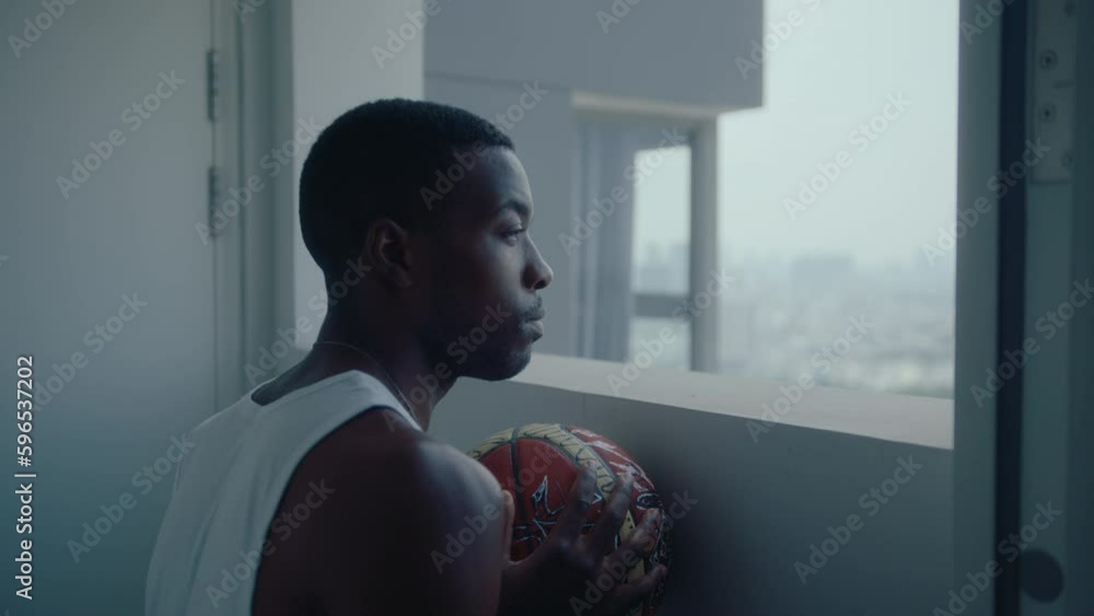 Young black basketball player holding a ball, looking pensively at the city through the window at the end of the hallway in an apartment building. Rack focus shot Young black basketball player holding a ball, looking pensively at the city through the window at the end of the hallway in an apartment building. Rack focus shot