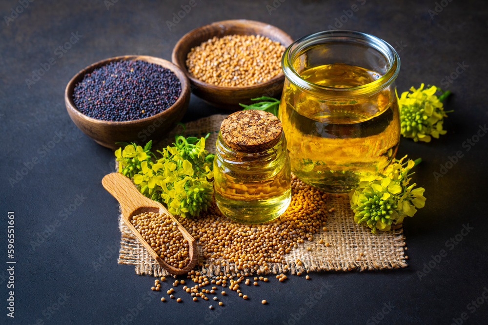 Mustard oil in a glass jar and decanter, mustard grains on a burlap napkin, flowers and leaves on wooden board background