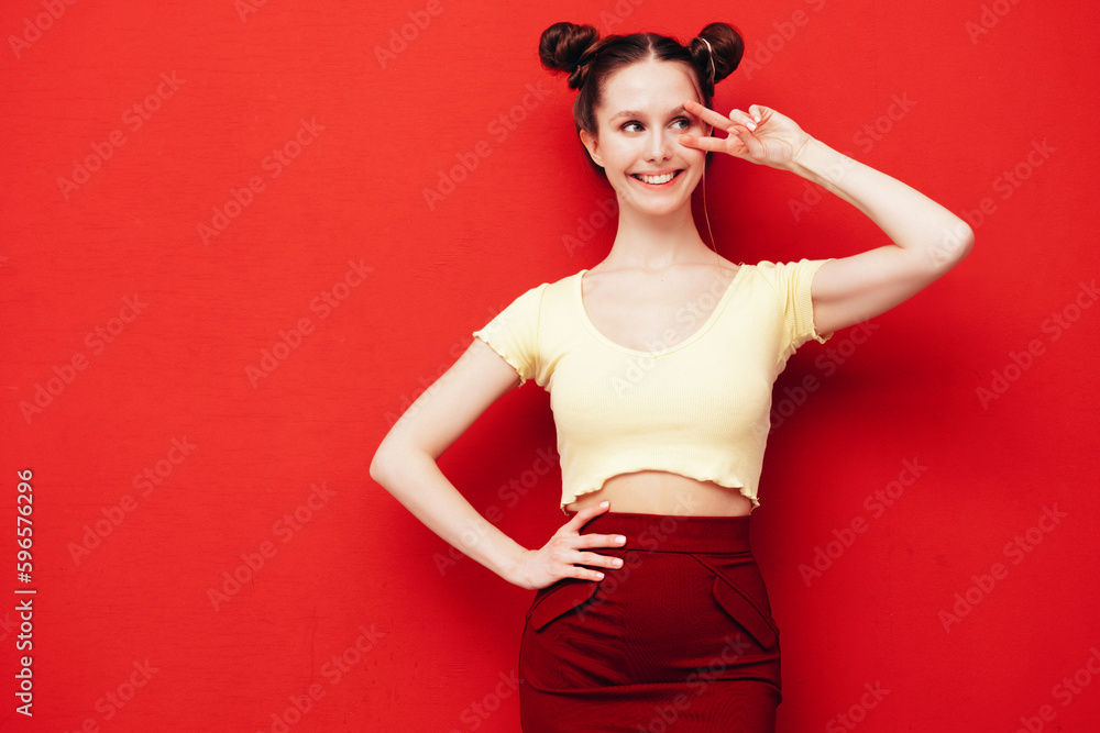 Young beautiful smiling female in trendy summer yellow t-shirt and skirt. Carefree woman with two horns hairstyle posing near red wall in studio. Positive model having fun. Shows peace sign Young beautiful smiling female in trendy summer yellow t-shirt and skirt. Carefree woman with two horns hairstyle posing near red wall in studio. Positive model having fun. Shows peace sign