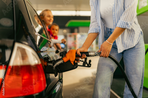 A young mother fills up gas tank at a gas station while her daughters look out the window