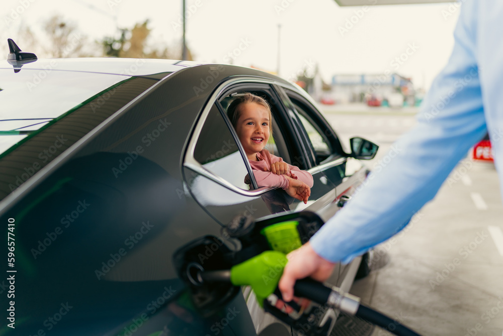 Fototapeta premium The daughter sits in the car leaning against the window and watches her father pour fuel into the tank