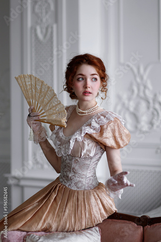 Young red haired woman in ball historic dress and fan in hand in vintage room sitting on sofa. Portrait of aristicratic woman in historically decorated room. photo
