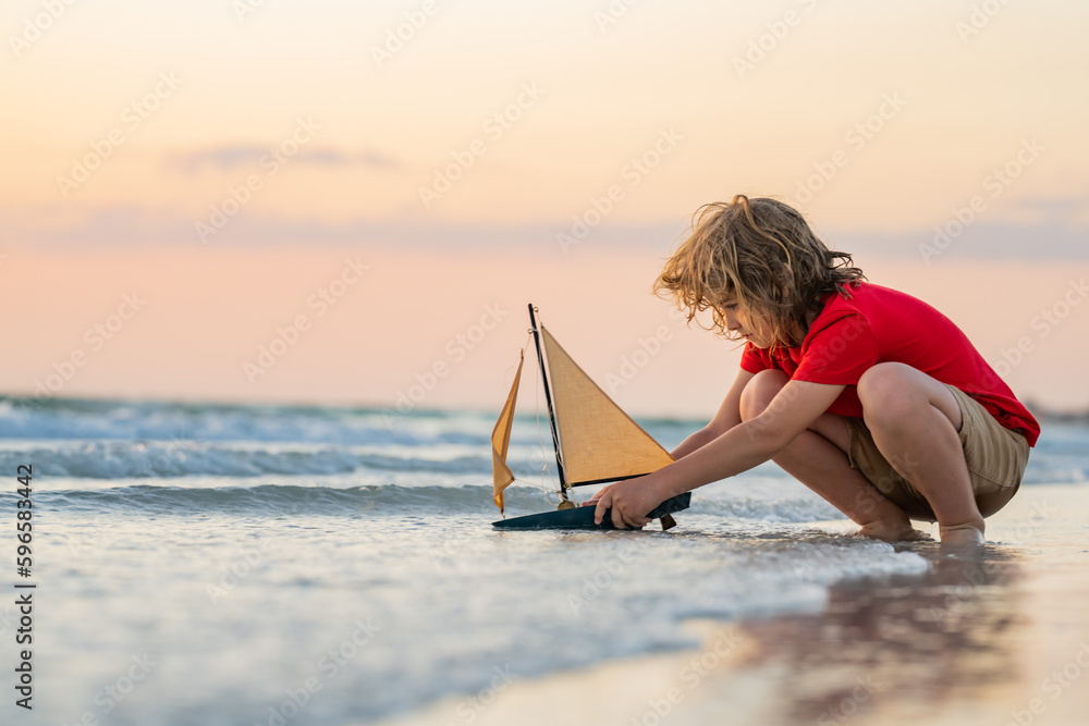 Kid boy playing with toy boat in sea water. Happy holiday by the sea. Kid dreaming about sailing. Happy kid playing with toy sailing boat. Summer vacation. Cute little kid hold toy sailboat. - obrazy, fototapety, plakaty 