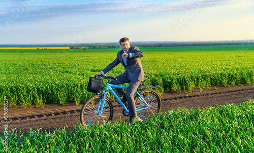 a businessman rides a bicycle with a briefcase and checks his wrist watch on a green grassy field, dressed in a business suit, beautiful nature in spring, freelance business concept