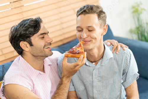 Cheerful gay couple eating pizza. Gay couple sitting on sofa at home, eating one slice of pizza. Homosexual relationship concept.