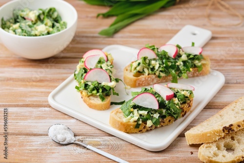 Open sandwiches with spring salad of wild garlic, boiled eggs and radish slices on a white ceramic board on a wooden background. Using wild plants for food,
