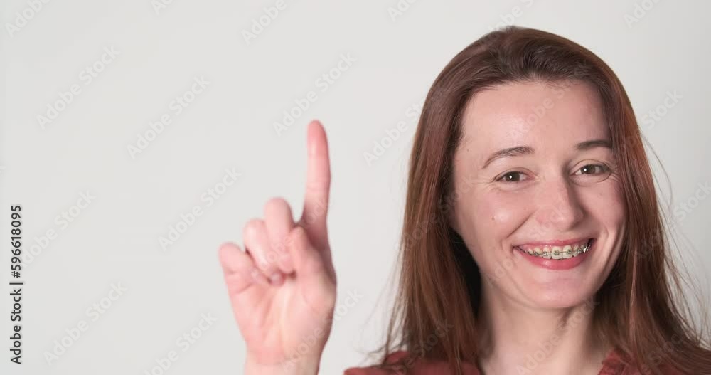 Beautiful young woman with braces on her teeth smiles and shows her index finger up. Gesture attention, close-up, portrait. Beautiful young woman with braces on her teeth smiles and shows her index finger up. Gesture attention, close-up, portrait.