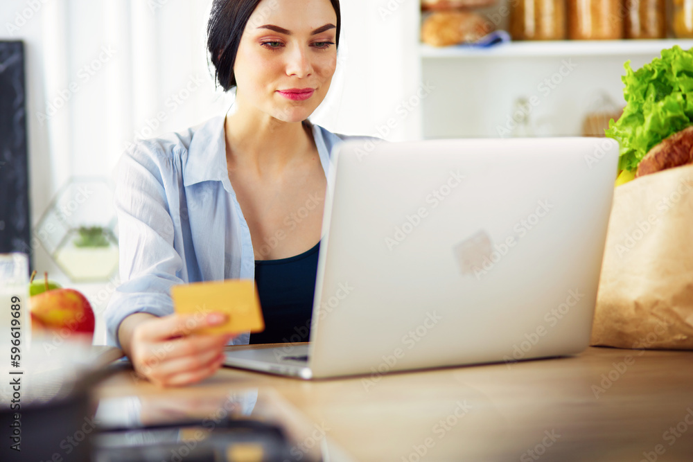 Smiling woman online shopping using computer and credit card in kitchen. Smiling woman online shopping using computer and credit card in kitchen.