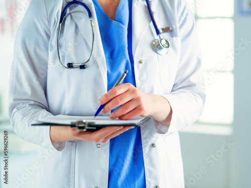 Doctor with a stethoscope, holding a notebook in his hand. Close-up of a female doctor filling up medical form at clipboard while standing straight in hospitalDoctor with a stethoscope, holding a note © lenets_tan