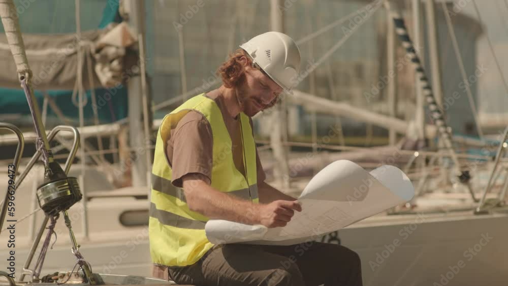 Medium shot of young Caucasian boat engineer in hard hat and acid green vest examining blueprint while sitting on sunny dock Medium shot of young Caucasian boat engineer in hard hat and acid green vest examining blueprint while sitting on sunny dock