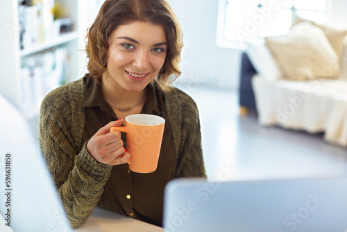 Attractive smiling woman sitting at office desk, holding a cup of coffee, she is relaxing and looking away.