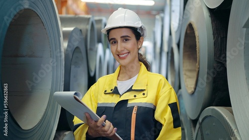 Hispanic latina woman industrial worker in uniform and helmet holding clipboard smiling and standing at industry manufacturing steel metal factory looking at camera