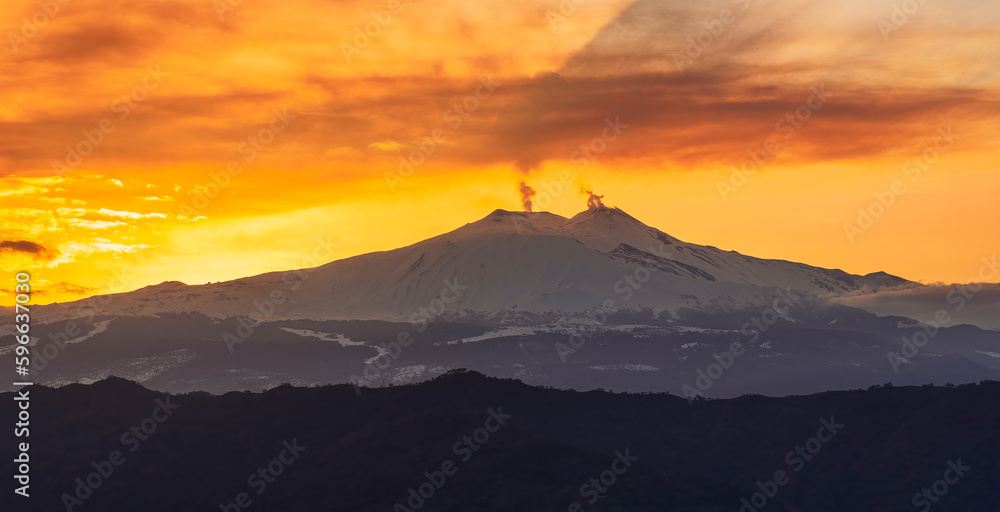 mysterious landscape of great erupting volcano with smoke from craters and snow on slopes in orange light of sunset. eruption of vulcan
