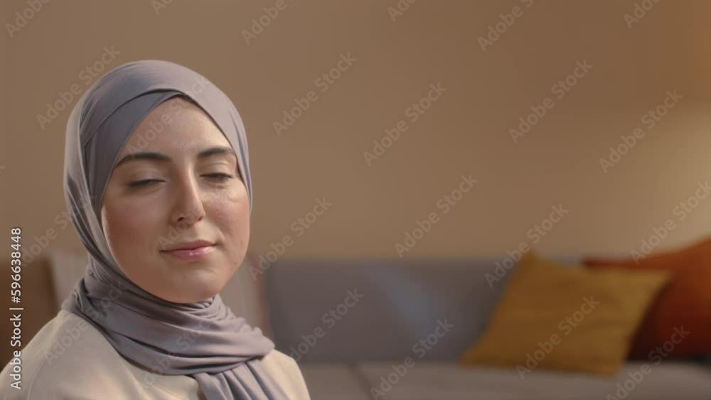 Chest up portrait of young smiling Muslim woman in hijab posing for camera in cozy living room