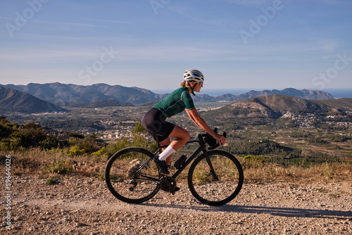 Fit female cyclist riding a gravel bike on a gravel road with a view of the mountains Coll de Rates pass.Alicante, Spain