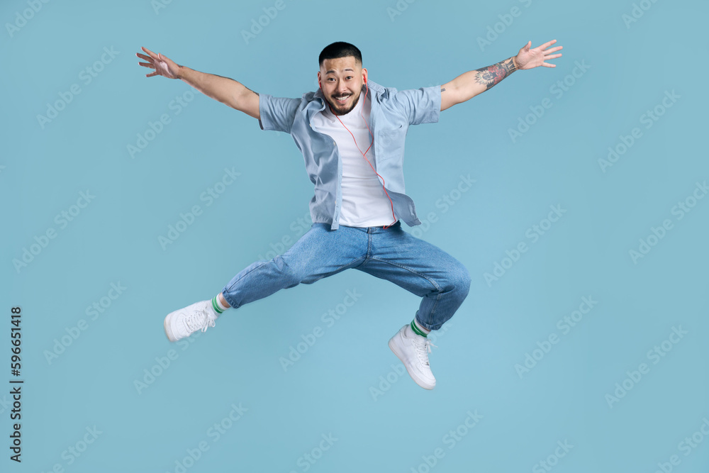 Happy asian man wearing  casual clothes, white shoes, stylish jeans jumping high isolated on blue background. Modern hipster listening music, positive lifestyle. Technology concept