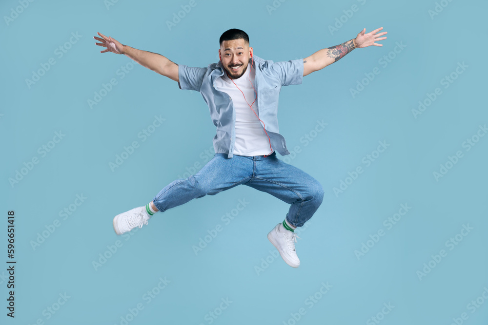 Happy asian man wearing casual clothes, white shoes, stylish jeans jumping high isolated on blue background. Modern hipster listening music, positive lifestyle. Technology concept Happy asian man wearing casual clothes, white shoes, stylish jeans jumping high isolated on blue background. Modern hipster listening music, positive lifestyle. Technology concept