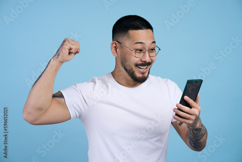 Fotografía Overjoyed asian man holding mobile phone sports betting, win money isolated on blue background
