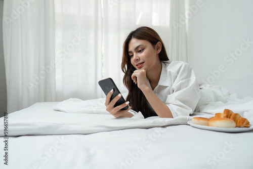 Young woman relaxing and eat bread using telephone on checking social apps and working the bedroom