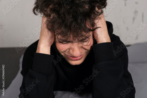 A severely depressed teenager, sitting on the bed, holding his head. A young man in a hard mental condition.