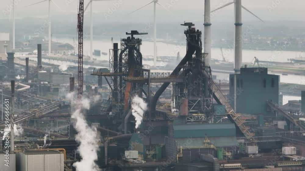 Aerial medium shot of Tata steel mill with wind turbines in the background, and one cutting through the frame Aerial medium shot of Tata steel mill with wind turbines in the background, and one cutting through the frame