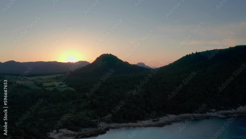 Drone capturing beautiful sunset at golden hour behind mountain hill with forest trees at the beach with calm clear blue water Drone capturing beautiful sunset at golden hour behind mountain hill with forest trees at the beach with calm clear blue water