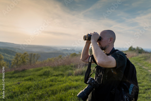 Adult young caucasian male traveler professional photographer looking through binoculars while walking in the mountains. Explore nature, trekking active lifestyle