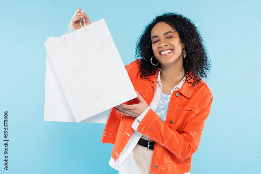 overjoyed african american woman in orange jacket holding white shopping bags isolated on blue. overjoyed african american woman in orange jacket holding white shopping bags isolated on blue.