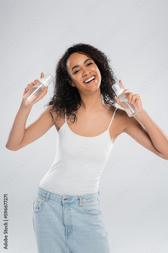 overjoyed african american woman in white tank top posing with bottles of face foam isolated on grey. overjoyed african american woman in white tank top posing with bottles of face foam isolated on grey.