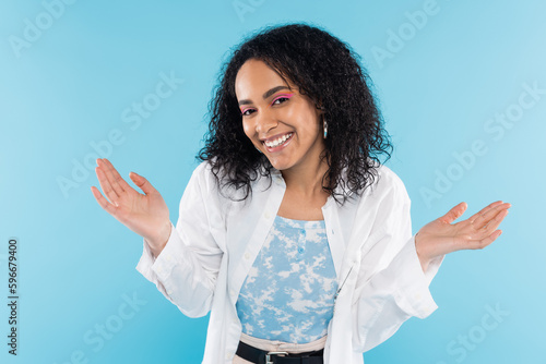cheerful african american woman looking at camera and showing shrug gesture isolated on blue.