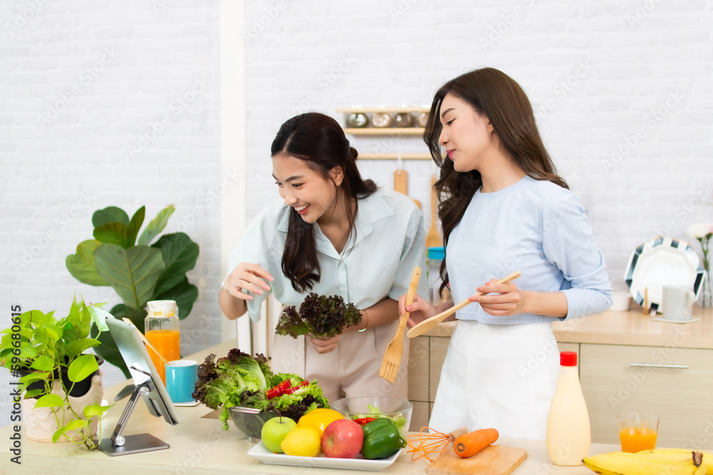 Two young Asian women are reading recipe from the digital tablet in the kitchen. Woman having fun with healthy food salad. Two young Asian women are reading recipe from the digital tablet in the kitchen. Woman having fun with healthy food salad.