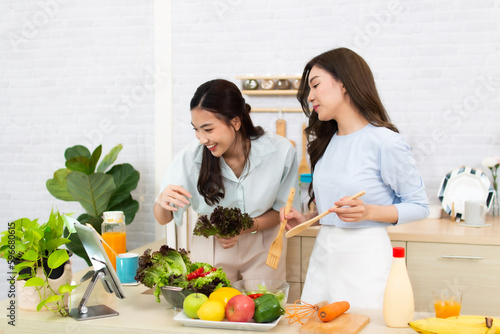Two young Asian women are reading recipe from the digital tablet in the kitchen. Woman having fun with healthy food salad.