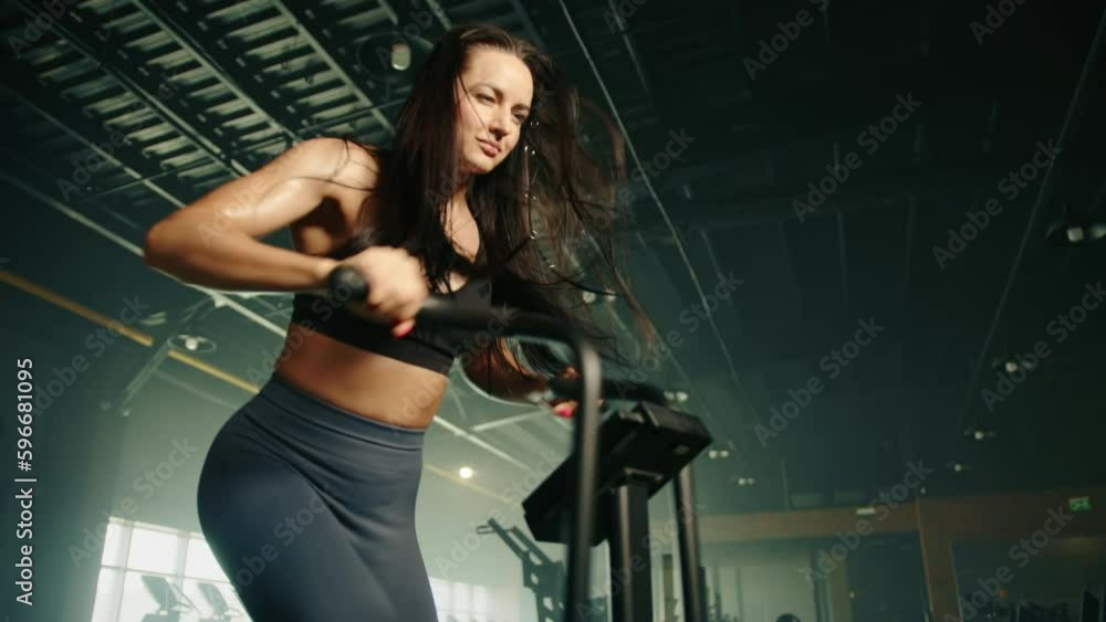 Medium close up of athletic young brown-haired woman exercising on air bike. Hair is wind blown. Dark ceiling, big window with daylight in the background. High quality 4k footage Medium close up of athletic young brown-haired woman exercising on air bike. Hair is wind blown. Dark ceiling, big window with daylight in the background. High quality 4k footage
