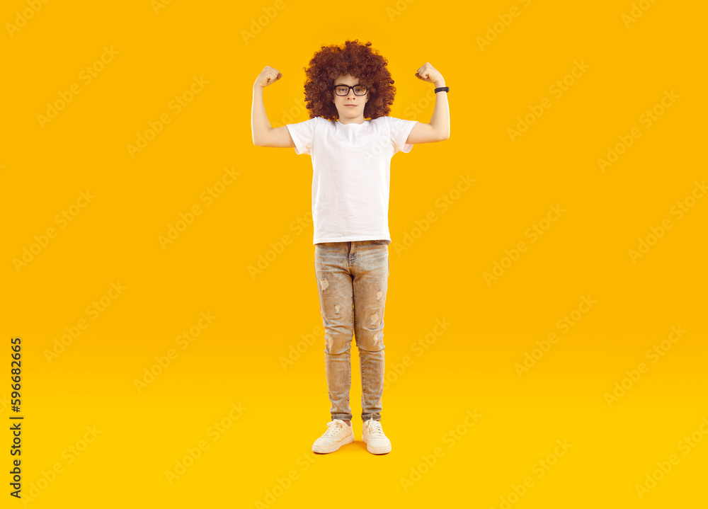 Funny child showing muscles. Little boy demonstrating his strength. Funny kid in a curly wig, white T shirt and glasses standing on a yellow background, pretending to be strong and flexing his arms Funny child showing muscles. Little boy demonstrating his strength. Funny kid in a curly wig, white T shirt and glasses standing on a yellow background, pretending to be strong and flexing his arms