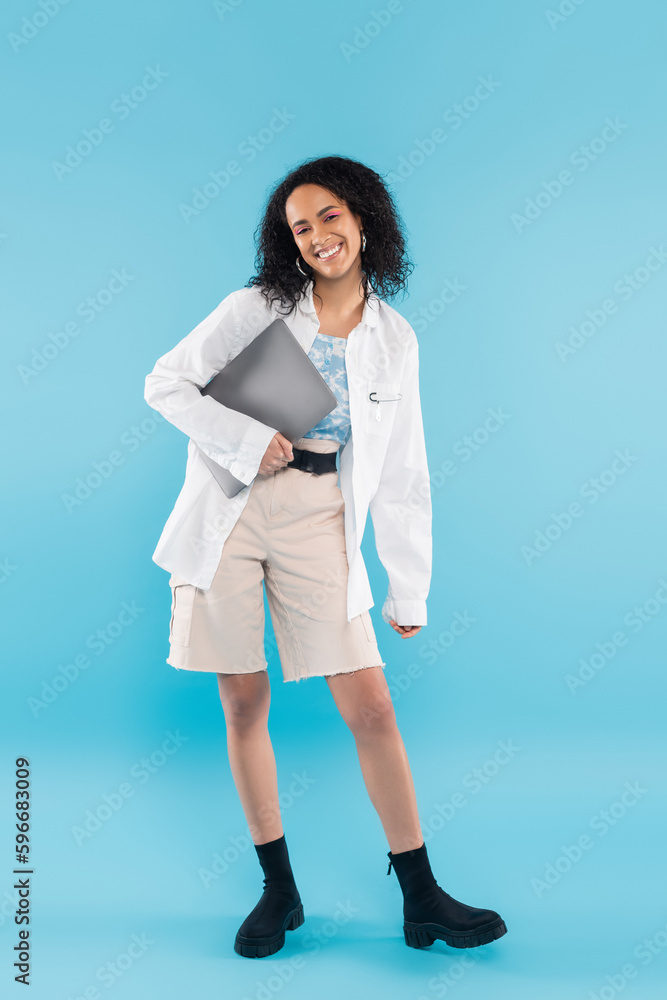 full length of carefree african american woman in white shorts and black boots posing with laptop on blue background. full length of carefree african american woman in white shorts and black boots posing with laptop on blue background.