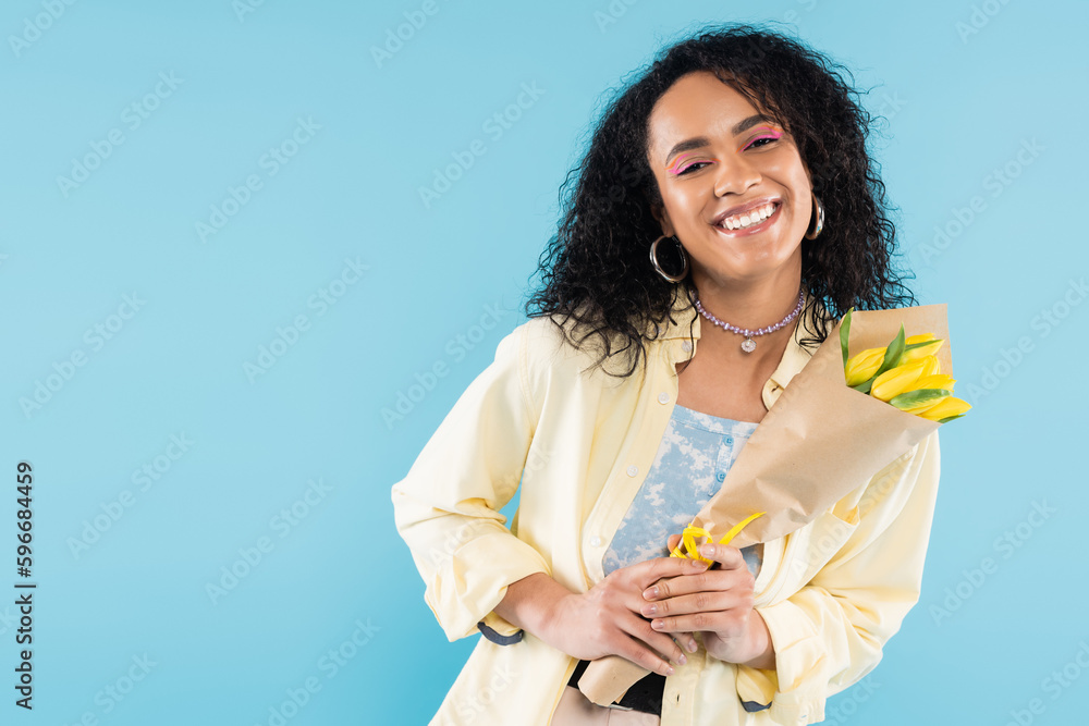 trendy african american woman in hoop earrings and beads holding yellow tulips and smiling at camera isolated on blue. trendy african american woman in hoop earrings and beads holding yellow tulips and smiling at camera isolated on blue.