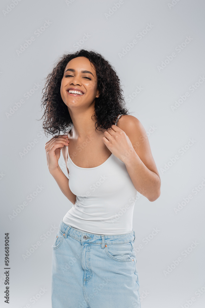 overjoyed african american woman in white tank top smiling at camera isolated on grey. overjoyed african american woman in white tank top smiling at camera isolated on grey.
