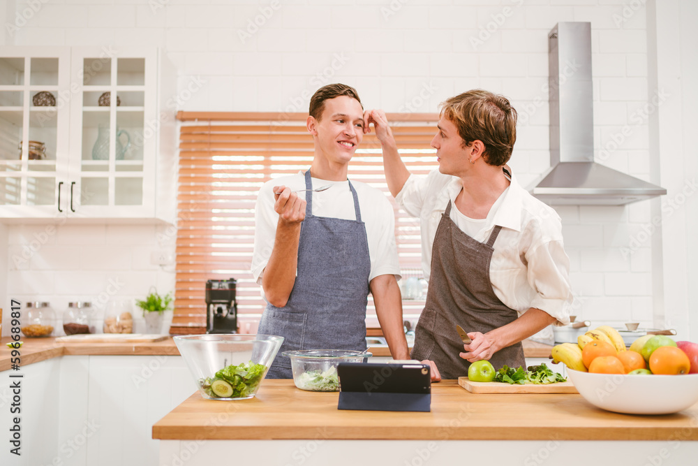 Portrait of the happy caucasian gay couple wearing aprons, looking at each other with smiles and enjoying cooking together in the kitchen at home. LGBT relationships. Gay couple cooking concept. Portrait of the happy caucasian gay couple wearing aprons, looking at each other with smiles and enjoying cooking together in the kitchen at home. LGBT relationships. Gay couple cooking concept.