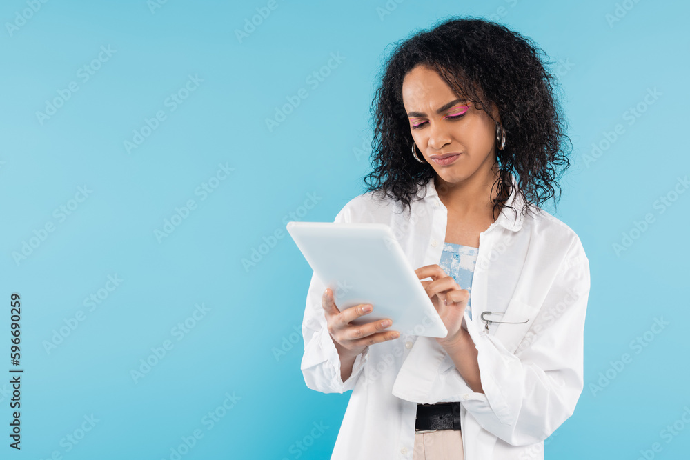displeased african american woman in white shirt using digital tablet isolated on blue. displeased african american woman in white shirt using digital tablet isolated on blue.