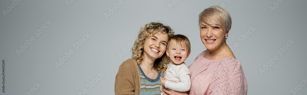excited baby girl laughing near happy mother and granny isolated on grey, banner. excited baby girl laughing near happy mother and granny isolated on grey, banner.