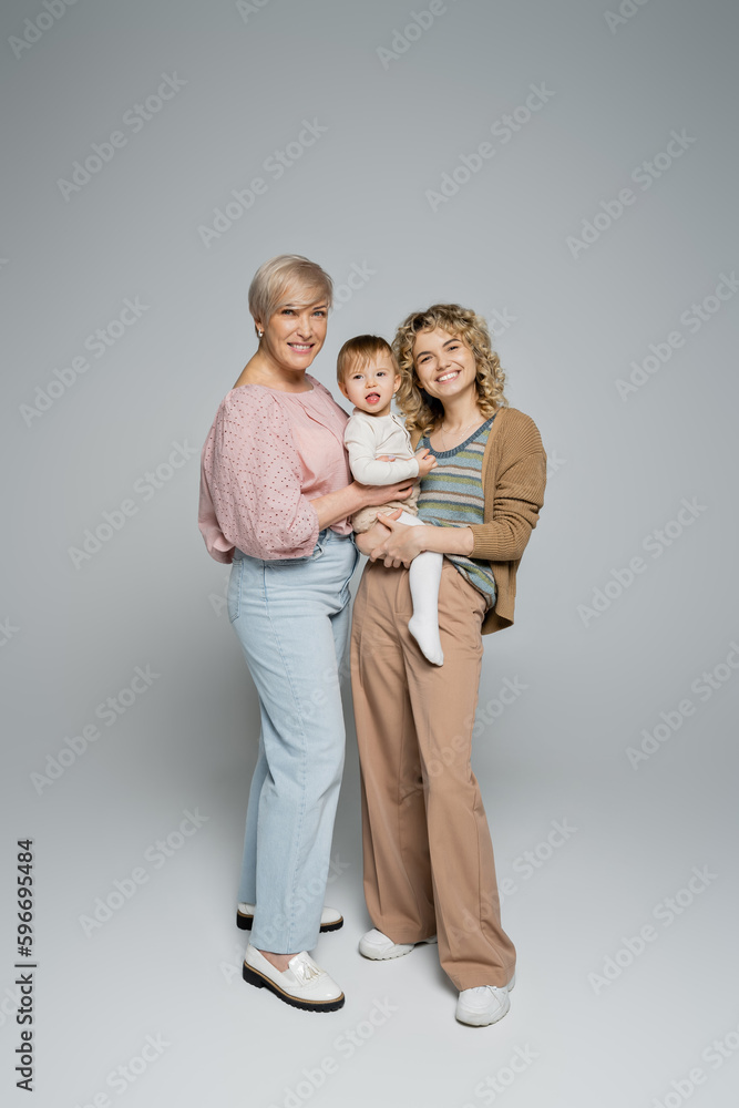 full length of mature woman smiling at camera near adult daughter with toddler child on grey background. full length of mature woman smiling at camera near adult daughter with toddler child on grey background.